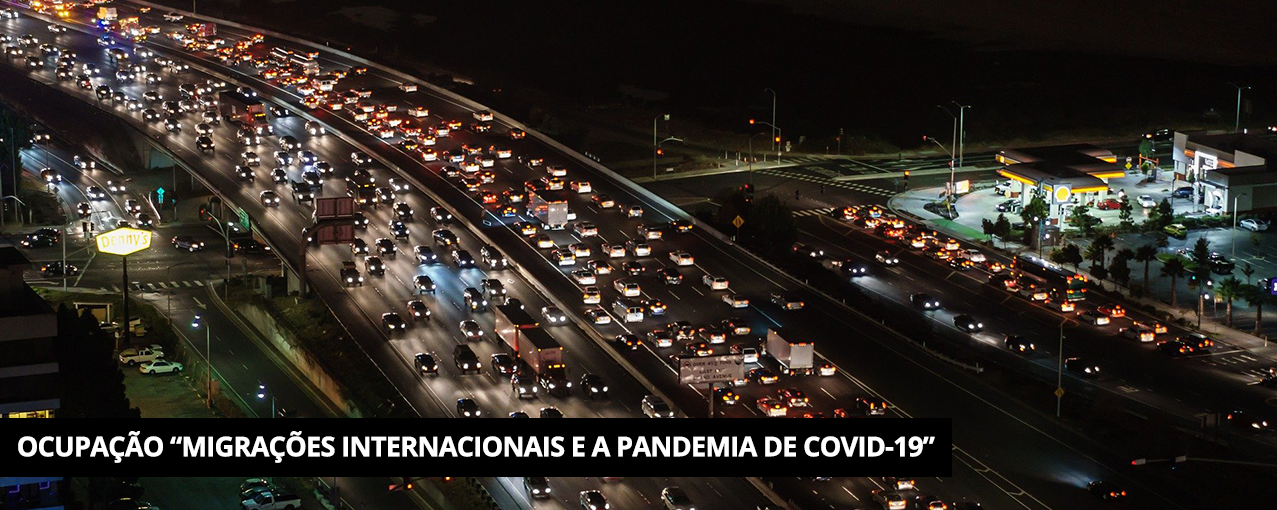 Colorida, de cima, mostrando, à noite, parte de uma cidade com ruas, viadutos e comércio. Tarja preta com Ocupação "Migrações Internacionais e a pandemia de COVID-19" escrito em branco